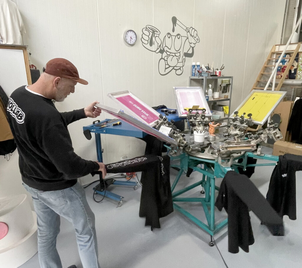 Studiofoto van Nollab, een creatieve werkruimte met zeefdrukmaterialen, verfblikken en gereedschap, waar kunstwerken en prints in verschillende stadia van productie zichtbaar zijn, met een rauwe, industriële sfeer en aandacht voor ambacht en detail