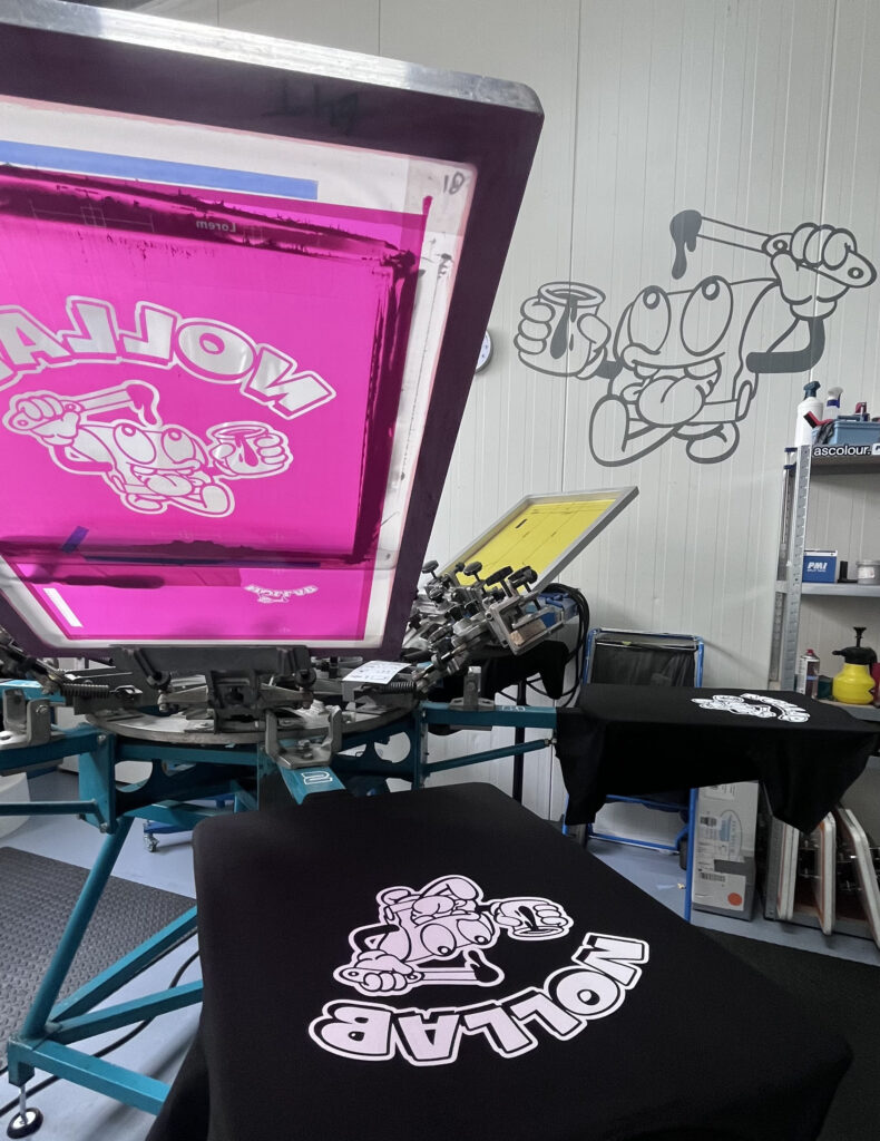 Studiofoto van Bollab, een creatieve werkruimte met zeefdrukmaterialen, verfblikken en gereedschap, waar kunstwerken en prints in verschillende stadia van productie zichtbaar zijn, met een rauwe, industriële sfeer en aandacht voor ambacht en detail